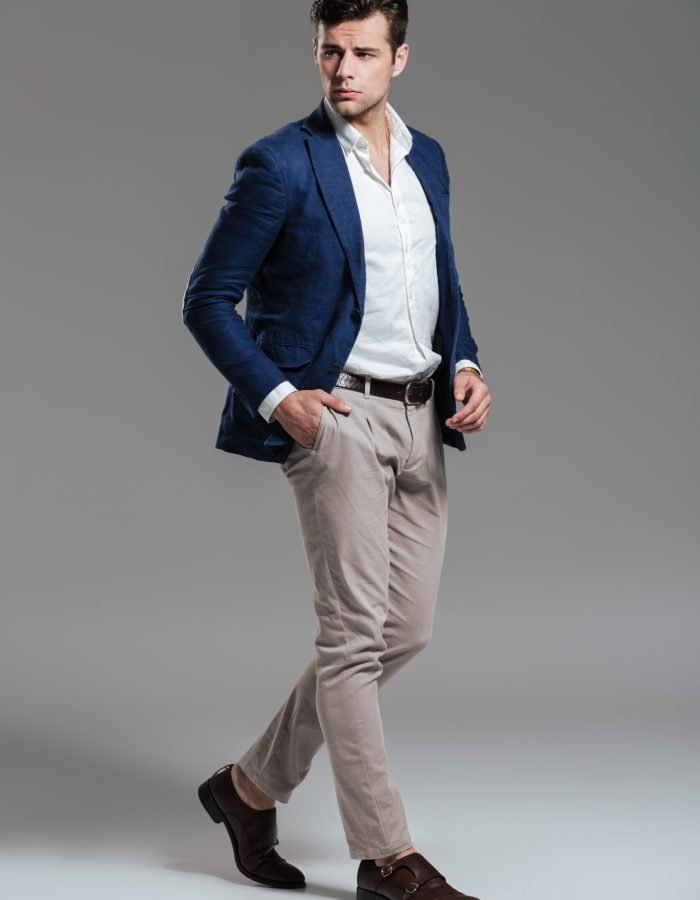 Full length portrait of a handsome serious man dressed in a jacket posing while standing and looking at camera isolated over gray background